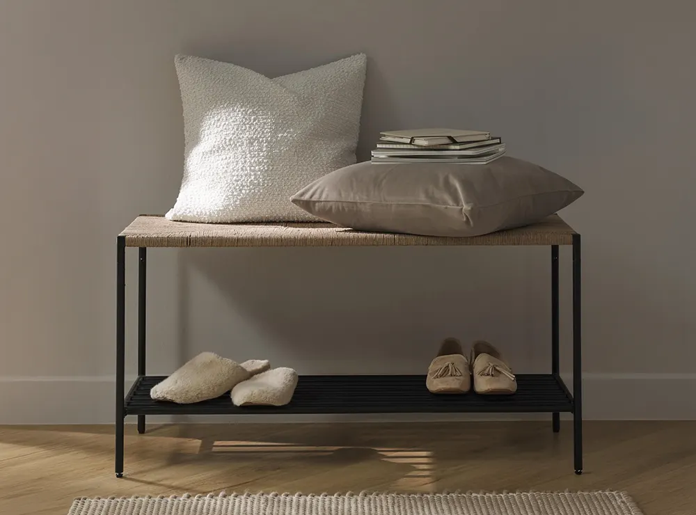 Weiße Bouclé-Kissenhülle und greiges Dekokissen auf einer geflochtenen Bank mit schwarzem Metallgestell, Materialvergleich für Sofa-Kissen