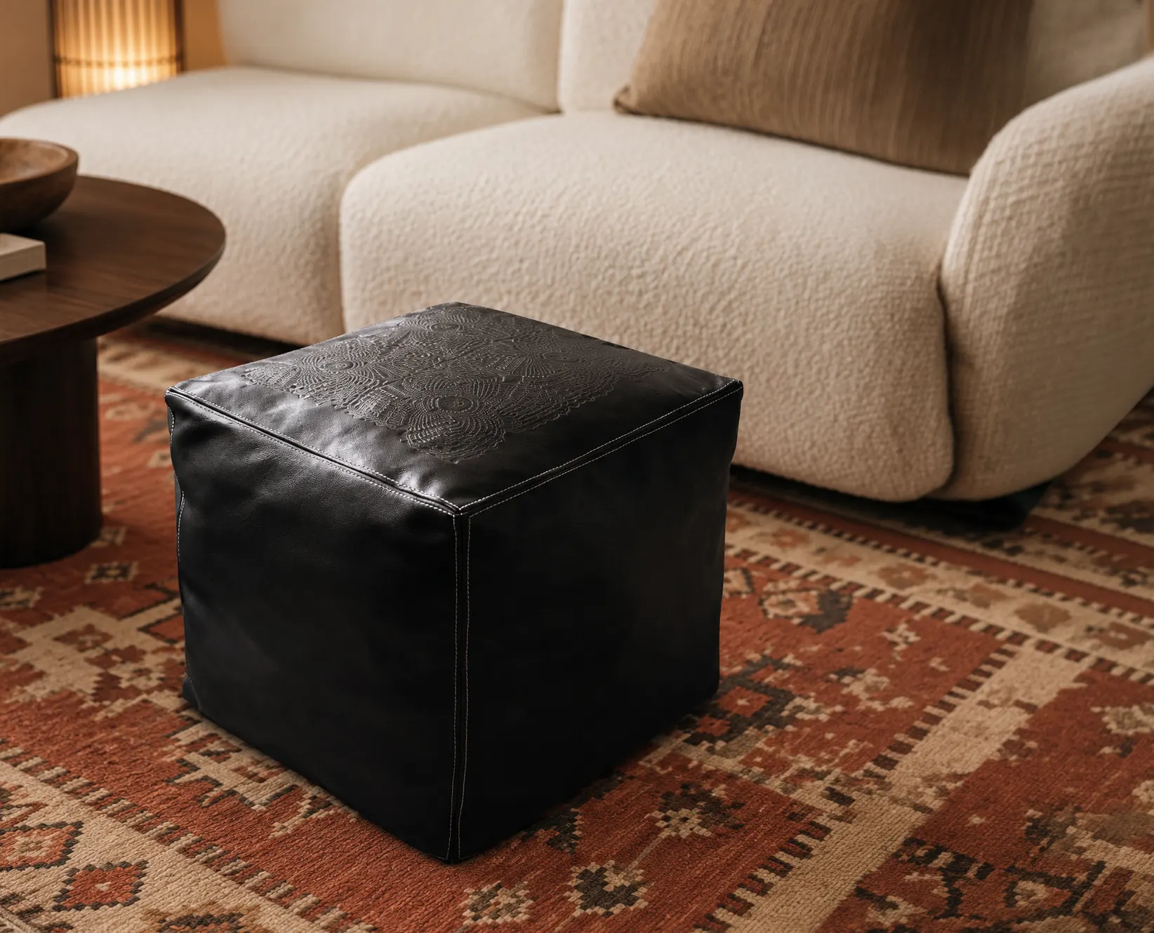 Quadratischer Leder-Pouf in Schwarz mit geprägtem Ornamentmuster auf Kelim-Teppich vor cremefarbenem Bouclé-Sofa