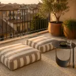 Low Lounge Balkon bei Goldstunde mit zwei Streifenkissen, schwarzem Metall-Beistelltisch und Olivenbaum vor mediterraner Stadtkulisse